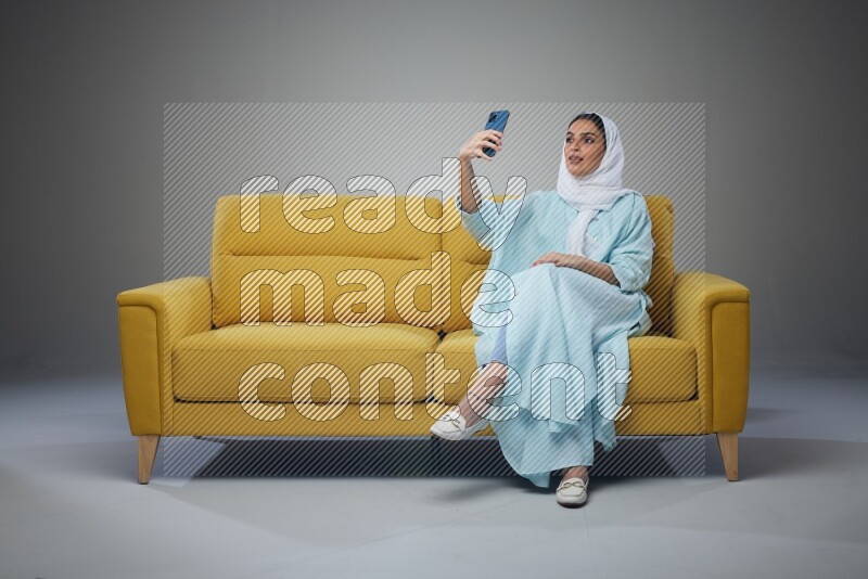 A Saudi woman wearing a light blue Abaya and white head scarf sitting on a yellow sofa and taking selfies with her phone eye level on a grey background