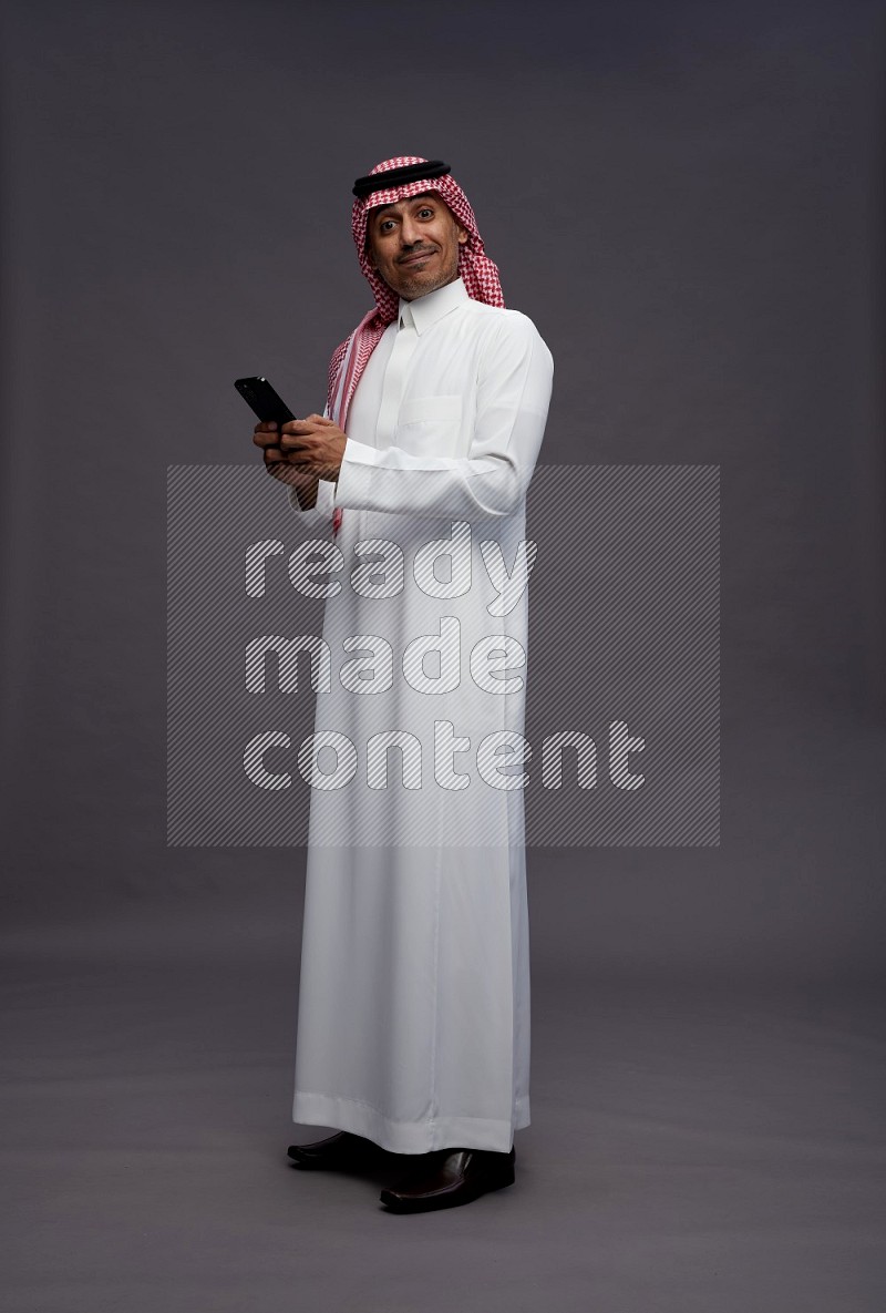 Saudi man wearing thob and shomag standing texting on phone on gray background