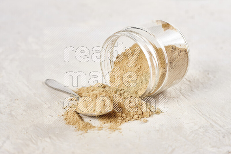 A glass jar full of ground ginger powder flipped with some spilling powder on white background