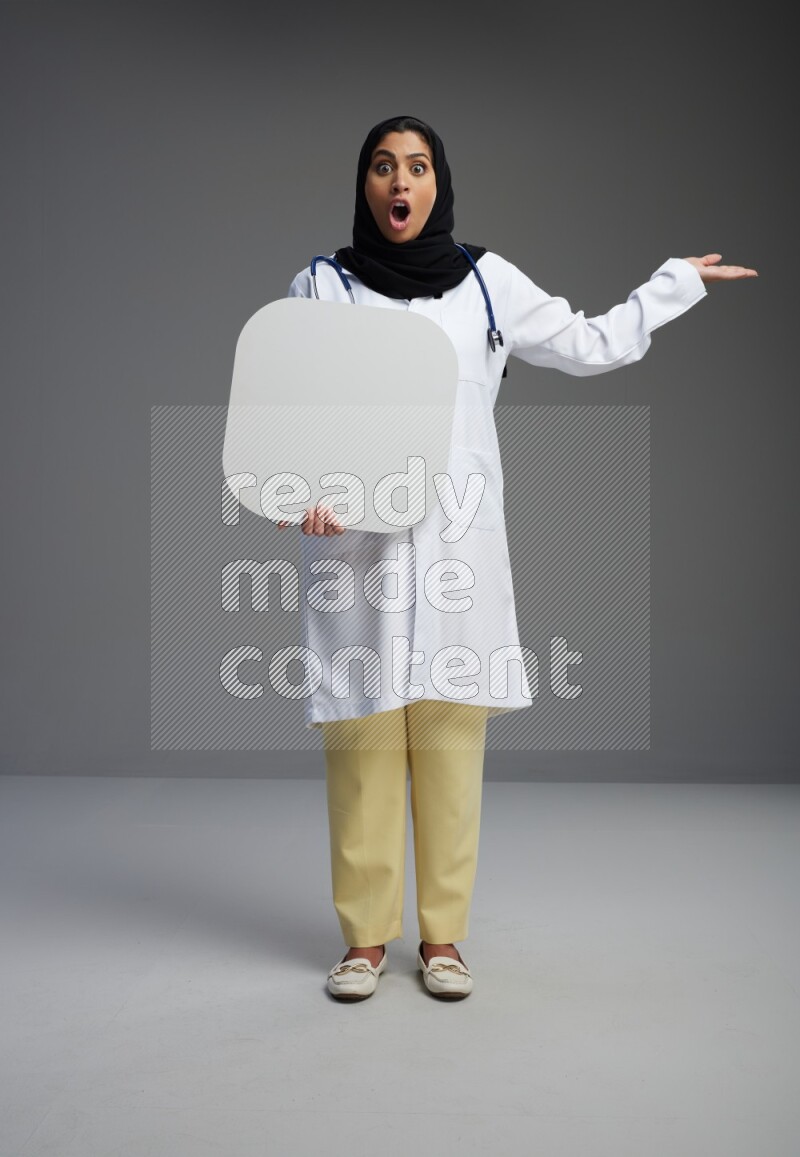 Saudi woman wearing lab coat with stethoscope standing holding social media sign on Gray background