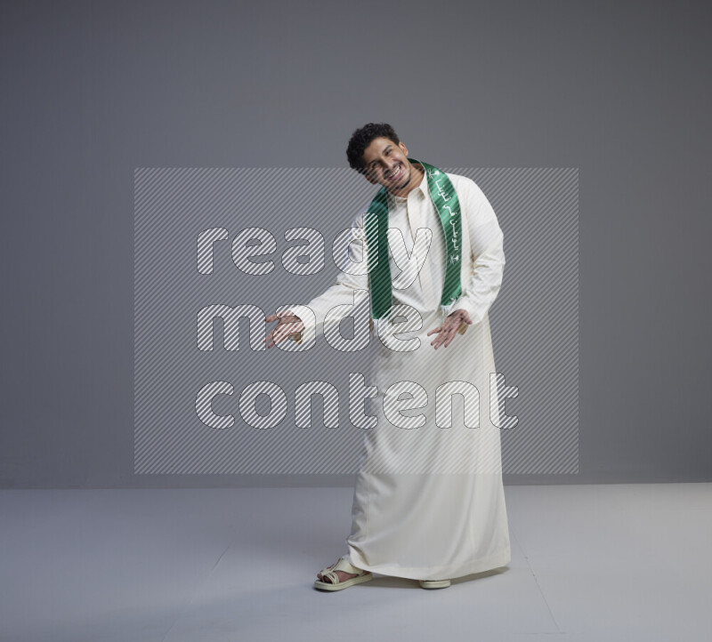 A Saudi man standing wearing thob and Saudi flag scarf on gray background