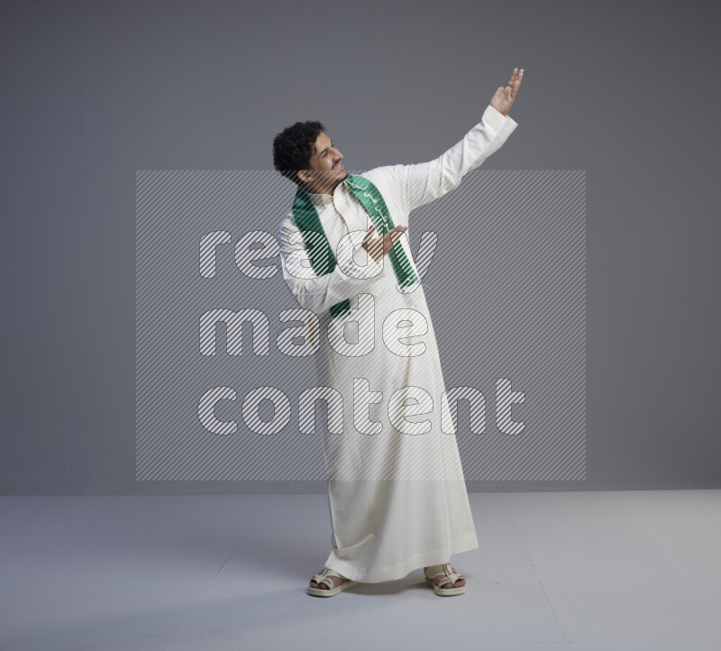 A Saudi man standing wearing thob and Saudi flag scarf on gray background