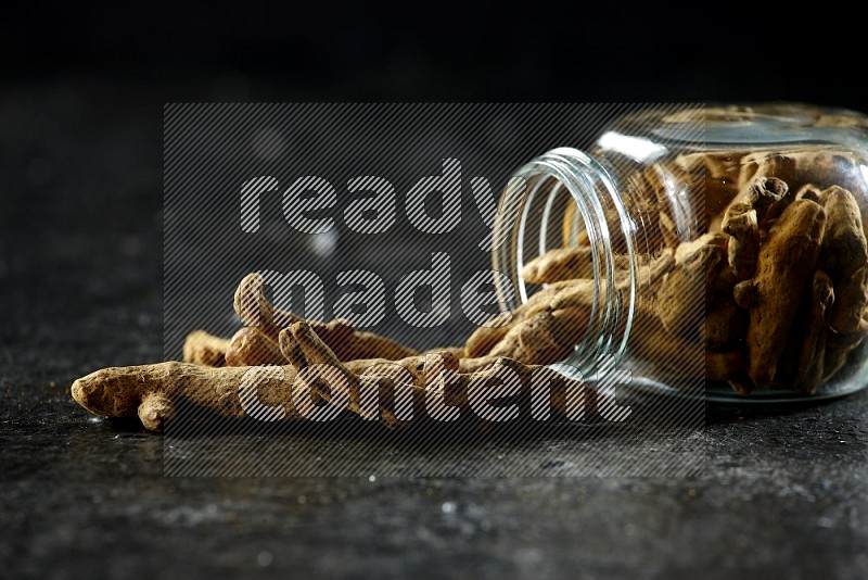 A flipped glass spice jar full of dried turmeric whole fingers and fingers fell out of it on textured black flooring