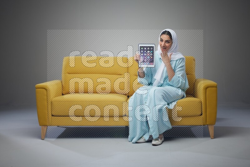 A Saudi woman wearing a light blue Abaya and white head scarf sitting on a yellow sofa and showing her tablet's screen while pointing to it eye level on a grey background