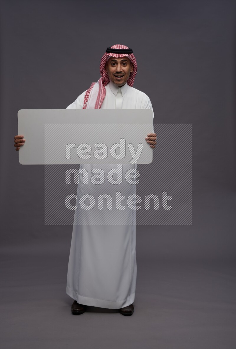 Saudi man wearing thob and shomag standing holding board on gray background