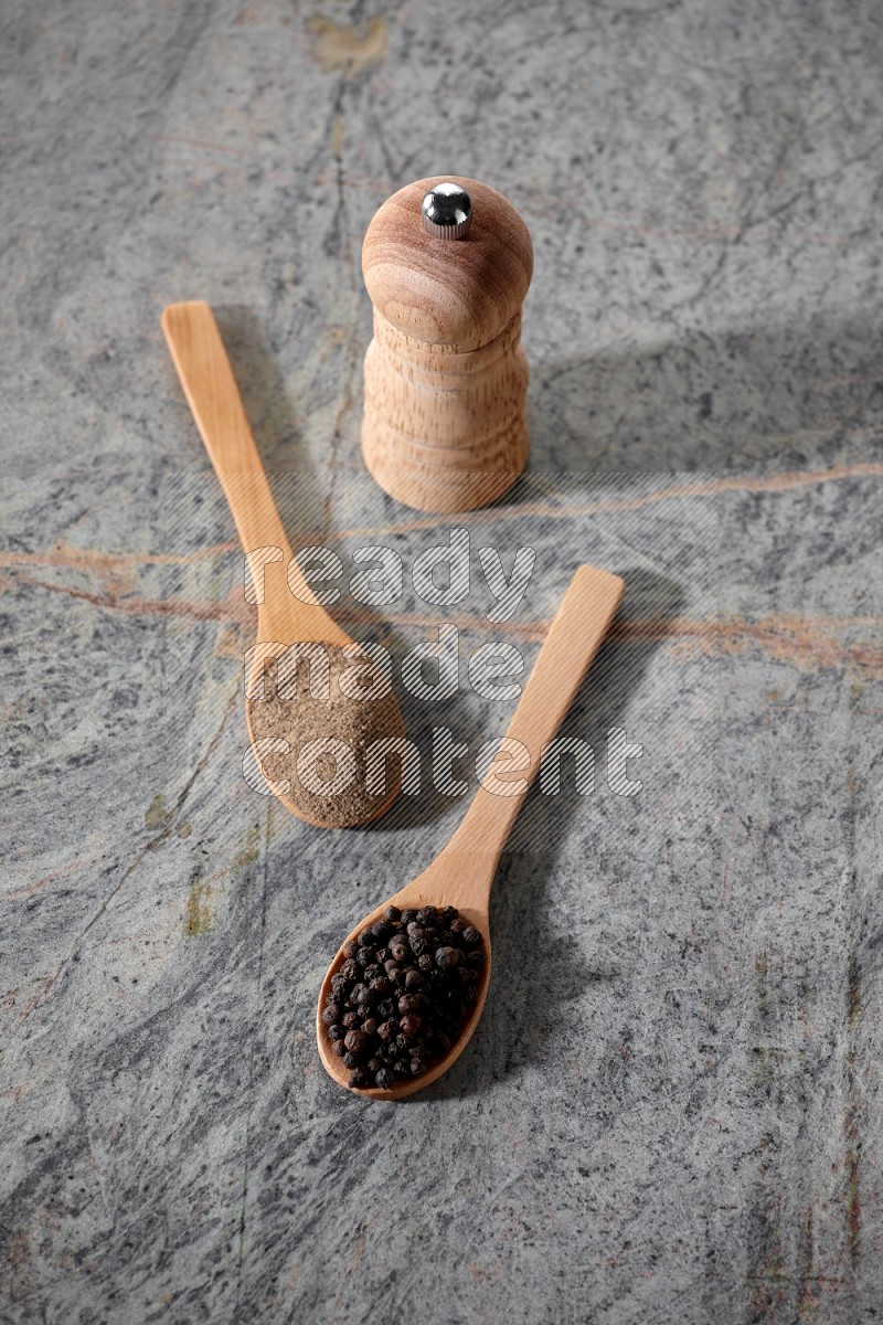 2 wooden spoons, one full of black pepper powder and the other full of black pepper beads and wooden grinder on a marble flooring
