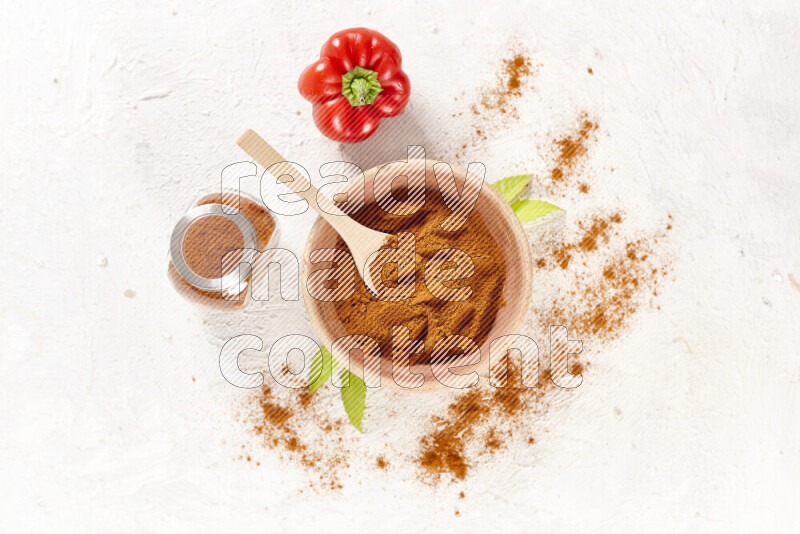 A wooden bowl full of ground paprika powder with a glass jar beside it and red bell pepper on white background