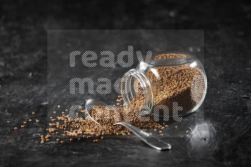 A glass spice jar and a metal spoon full of mustard seeds and jar is flipped with fallen seeds on a textured black flooring in different angles