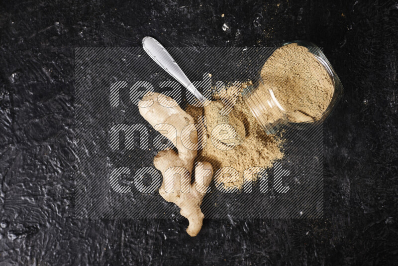 A glass jar full of ground ginger powder flipped with some spilling powder on black background
