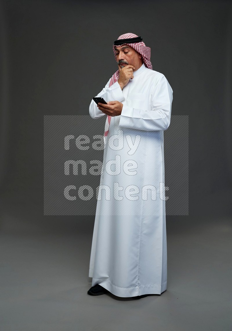 Saudi man with shomag Standing texting on phone on gray background