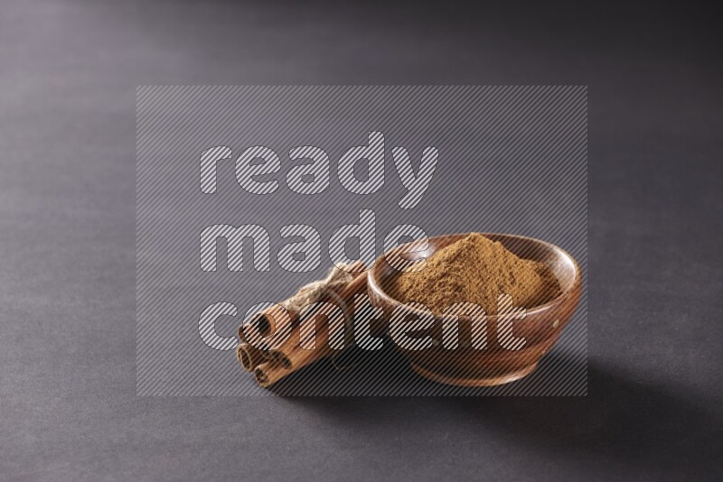 Cinnamon sticks stacked and bounded beside a wooden bowl full of cinnamon powder and a wooden spoon full of powder on black background