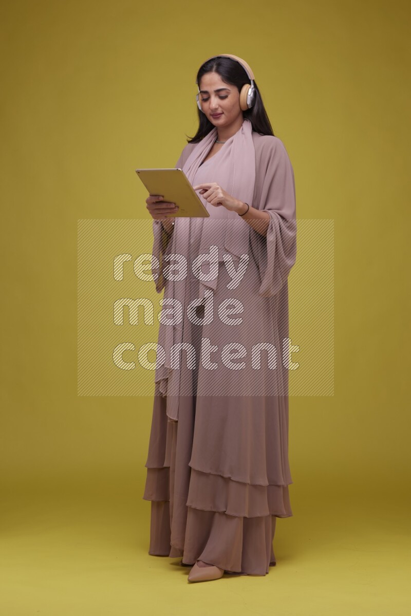 A woman Holding an Ipad with Headphone on a Yellow Background wearing Brown Abaya