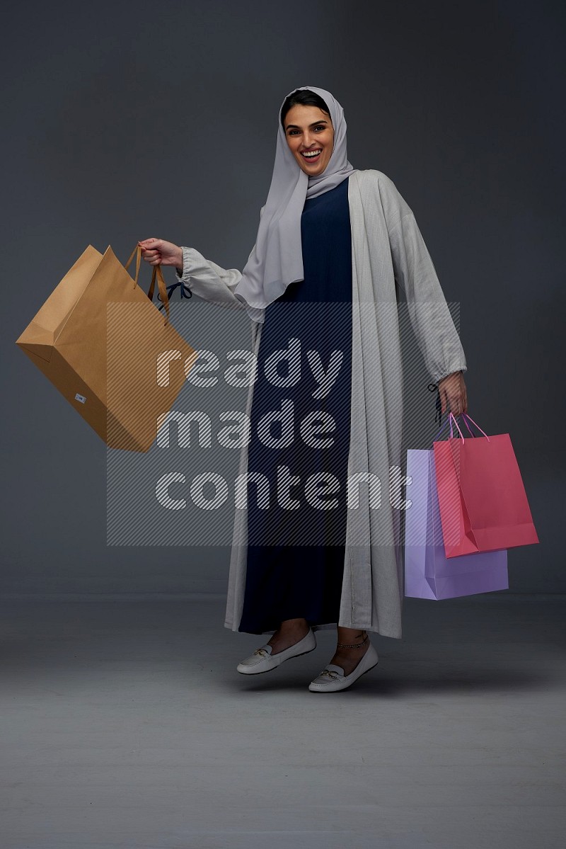 A Saudi woman wearing a light gray Abaya and head scarf standing and holding shopping bags on a grey background