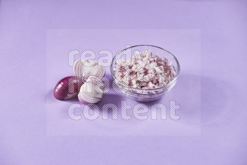 A glass bowl full of chopped red onions with halved onions beside it on purple background