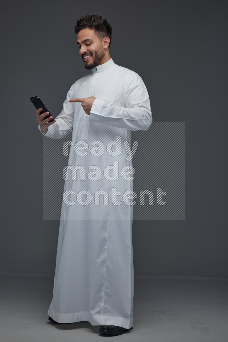 A Saudi man wearing Thobe and holding his phone while pointing to it eye level on a gray background