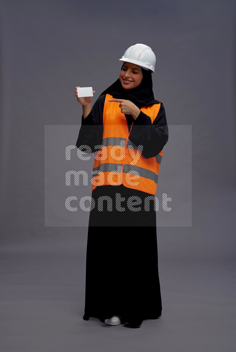 Saudi woman wearing Abaya with engineer vest standing holding ATM card on gray background