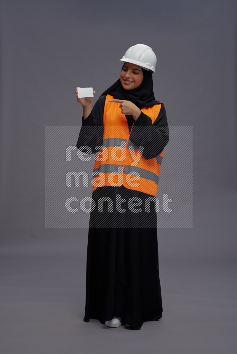 Saudi woman wearing Abaya with engineer vest standing holding ATM card on gray background