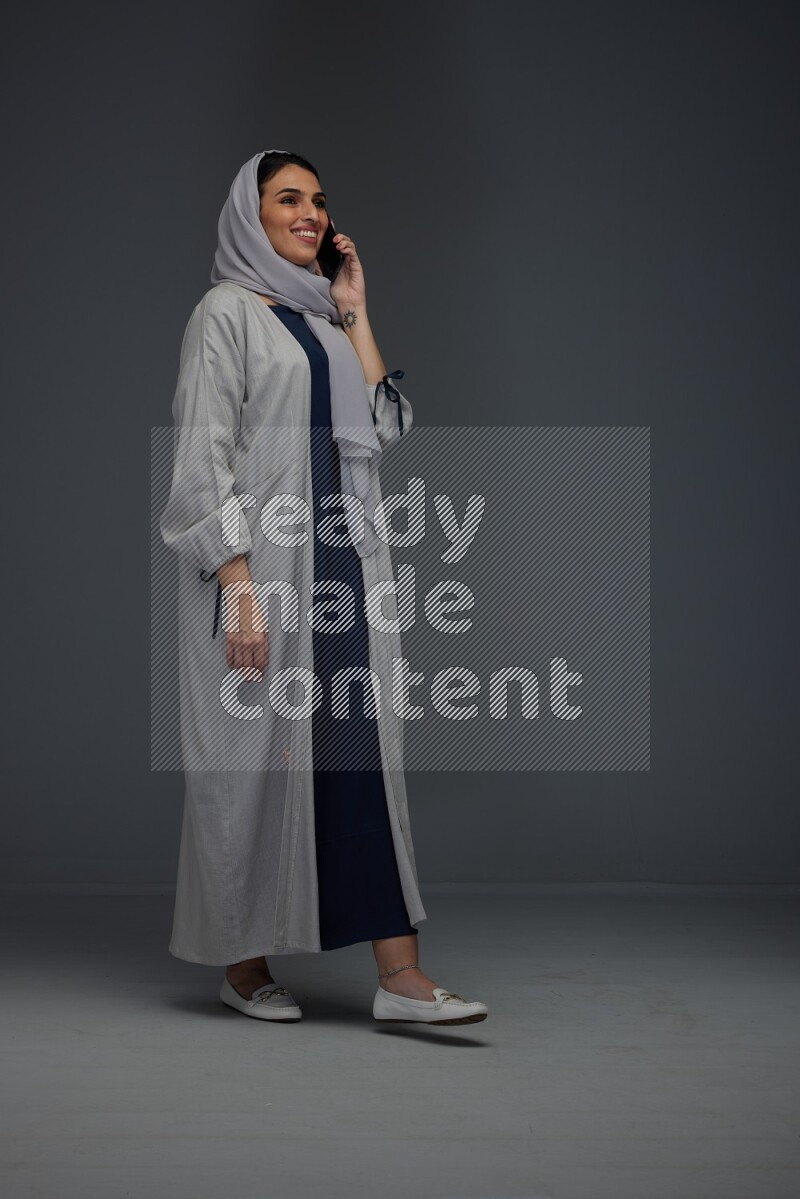 A Saudi woman wearing a light gray Abaya and head scarf standing and talking in the phone eye level on a grey background