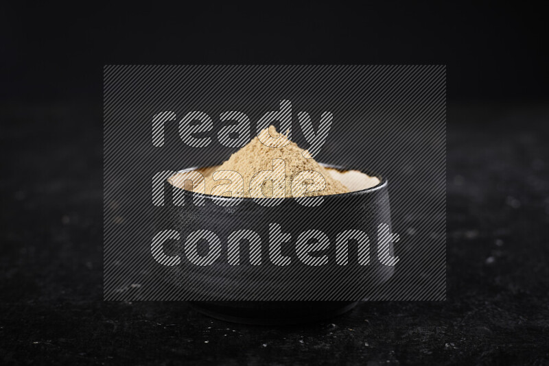 A black pottery bowl full of ground ginger powder on black background