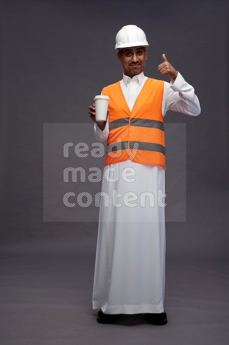 Saudi man wearing thob with engineer vest standing holding paper cup on gray background