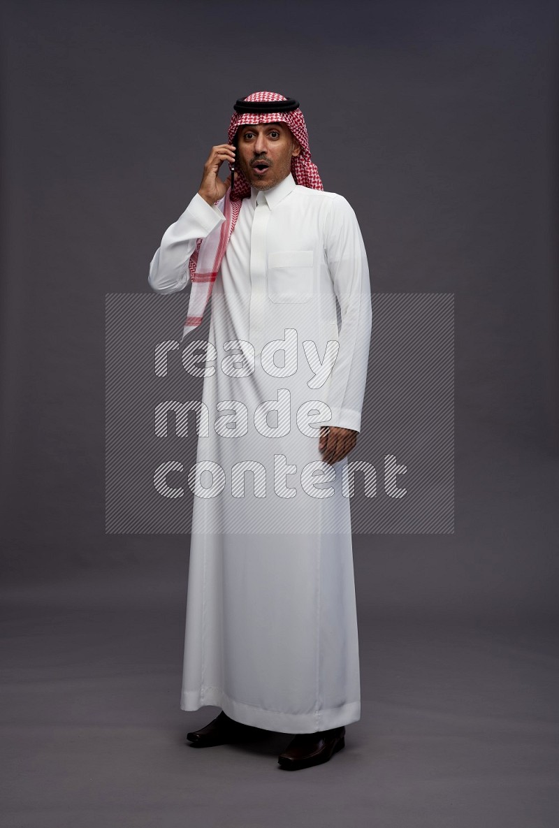 Saudi man wearing thob and shomag standing talking on phone on gray background
