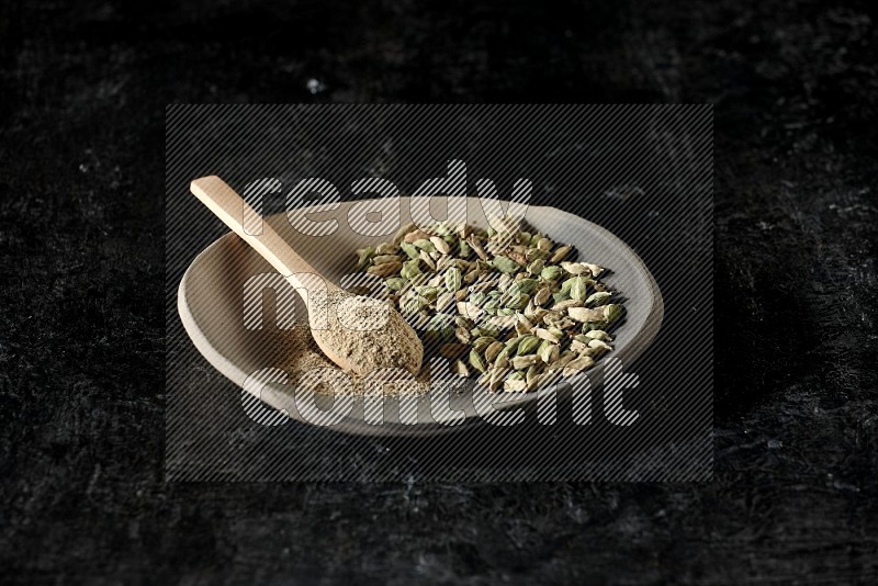 A plate filled with cardamom seeds and a wooden spoon full of cardamom powder on a textured black flooring
