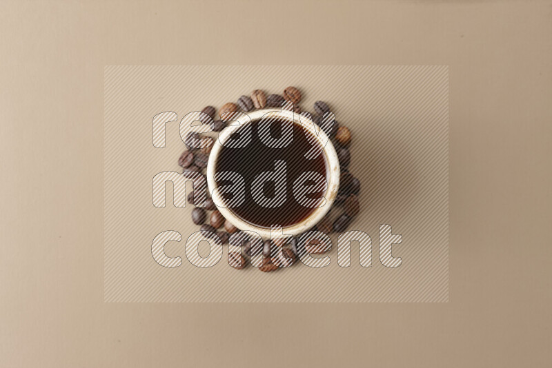 A beige pottery cup of coffee surrounded by roasted coffee beans on beige background