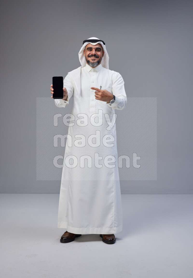 Saudi man Wearing Thob and white Shomag standing showing phone to camera on Gray background