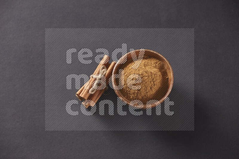 Cinnamon sticks stacked and bounded beside a wooden bowl full of cinnamon powder and a wooden spoon full of powder on black background