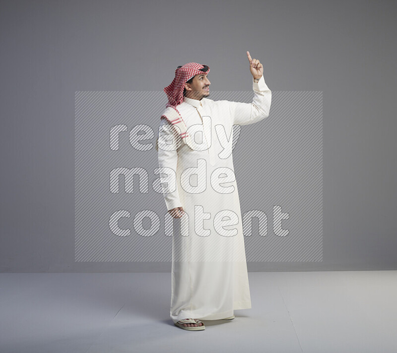 A Saudi man standing wearing thob and red shomag interacting with the camera on gray background