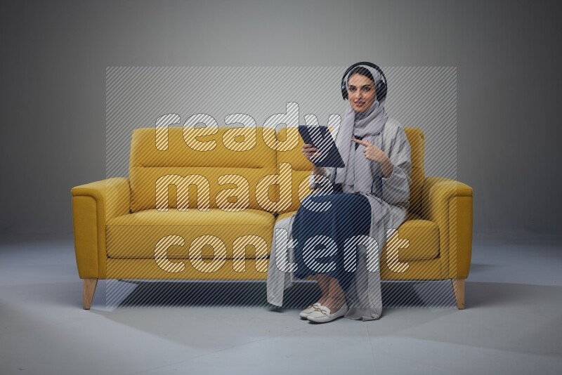 A Saudi woman wearing a light gray Abaya and head scarf sitting on a yellow sofa and using her phone while wearing headphones eye level on a grey background