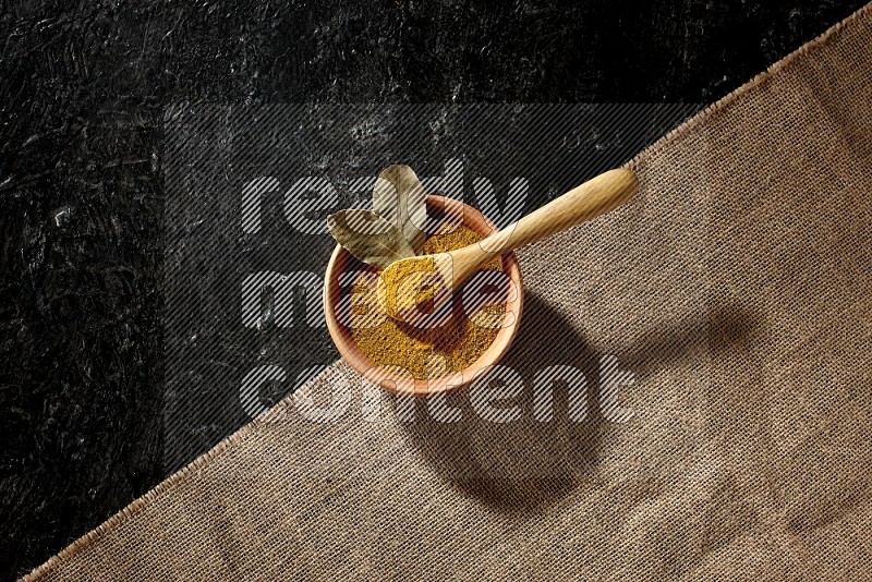 A wooden bowl and a wooden spoon full of turmeric powder on burlap fabric on textured black flooring