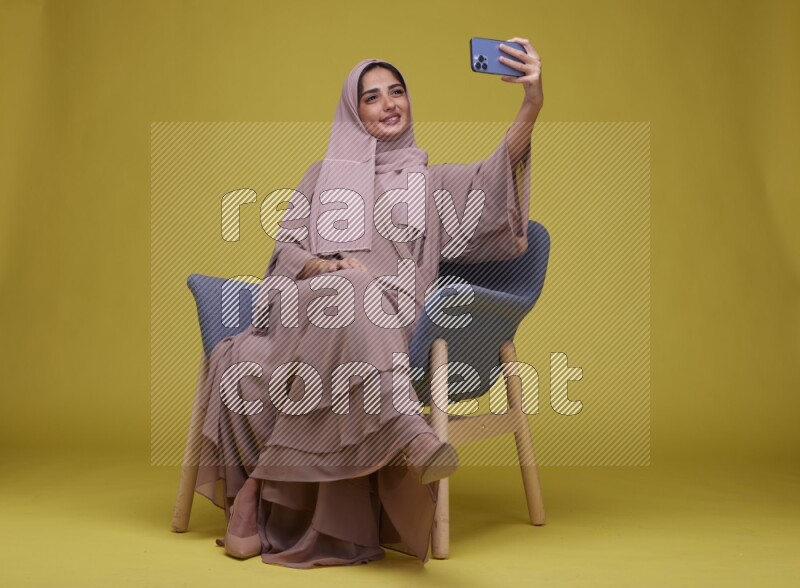 A woman sitting on a blue chair and Taking a Selfie  Setting  on a Yellow Background wearing Brown Abaya with Hijab