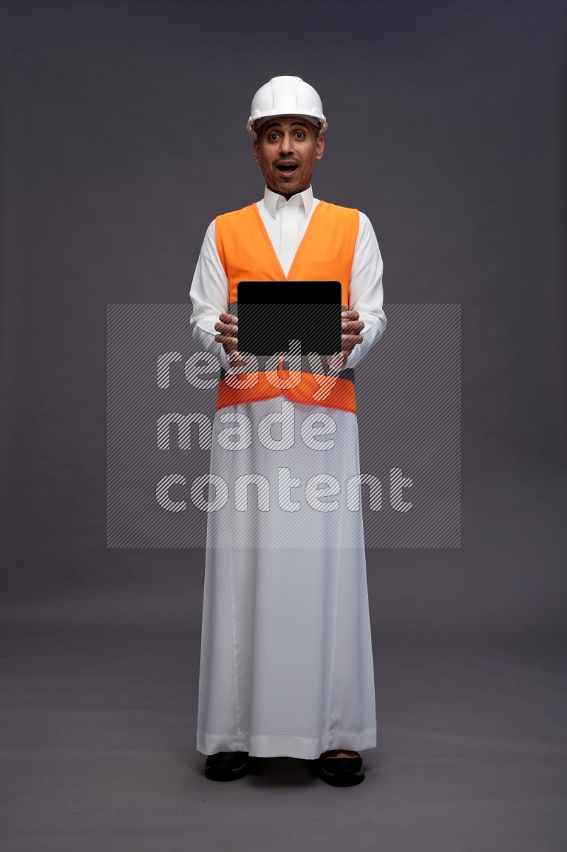 Saudi man wearing thob with engineer vest standing showing tablet to camera on gray background