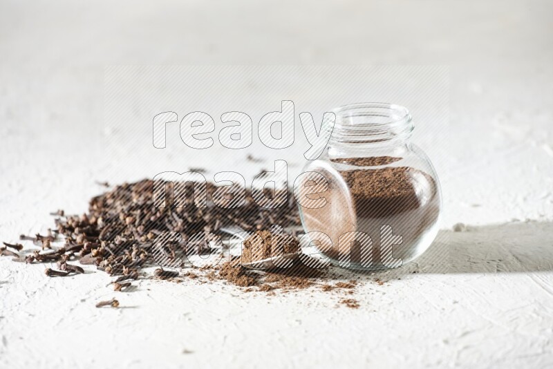 A glass spice jar and a metal spoon full of cloves powder and cloves spread on textured white flooring