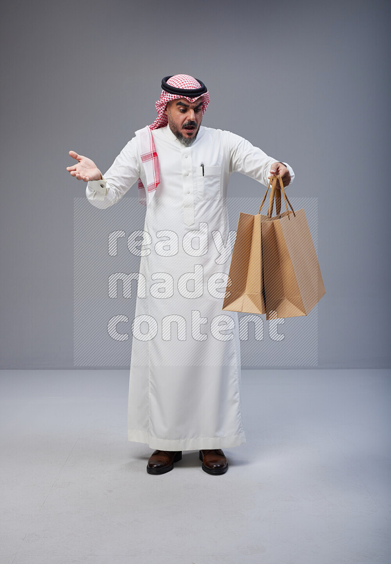 Saudi man Wearing Thob and red Shomag standing holding shopping bag on Gray background