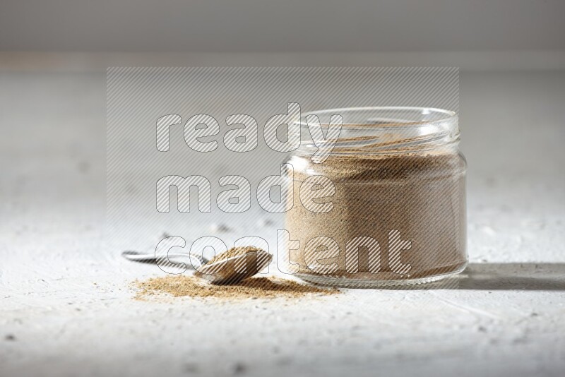 A glass jar and a metal spoon full of cumin powder on textured white flooring