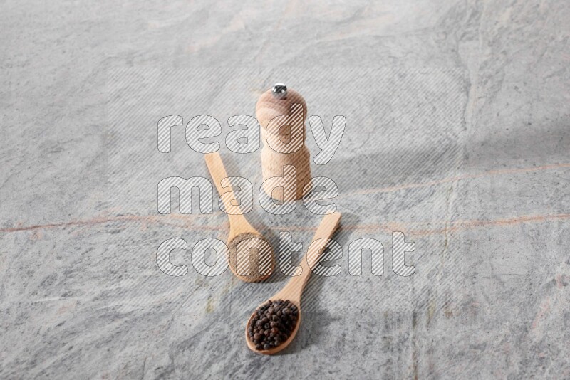 2 wooden spoons, one full of black pepper powder and the other full of black pepper beads and wooden grinder on a marble flooring