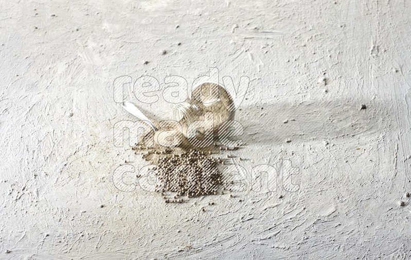A flipped herbal glass jar and metal spoon full of white pepper powder with spilled powder and beads on textured white flooring