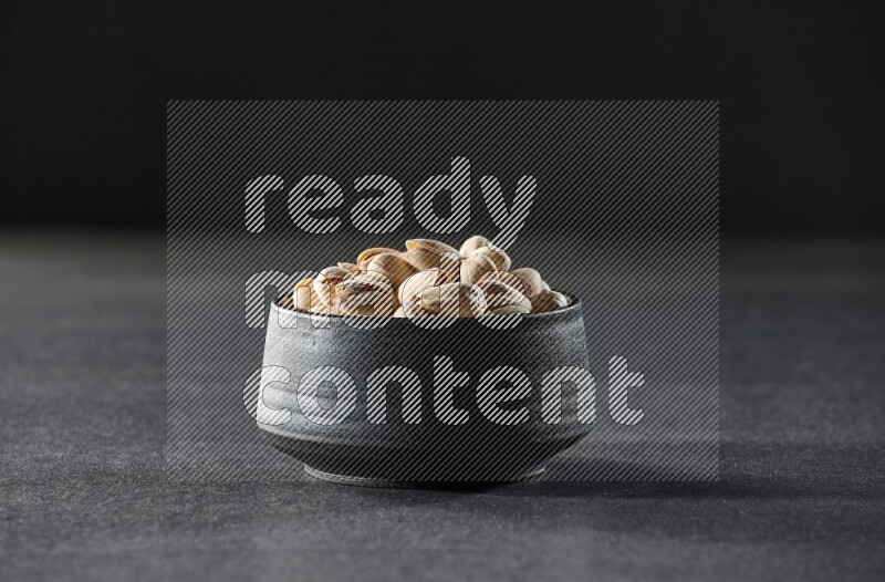 A black pottery bowl full of pistachios on a black background in different angles