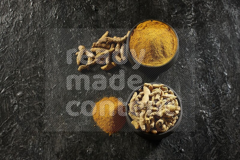 2 pottery black bowls, one full of turmeric powder and the other full of dried turmeric whole fingers on a textured black flooring