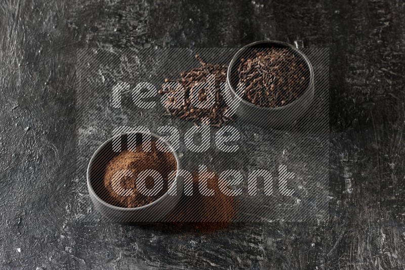 2 Black pottery bowls full of cloves and the other full of cloves powder on textured black flooring