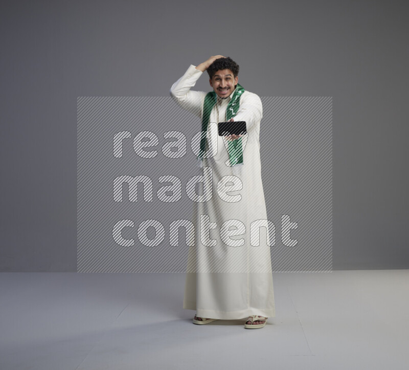 A Saudi man standing wearing thob and Saudi flag scarf showing phone to camera on gray background