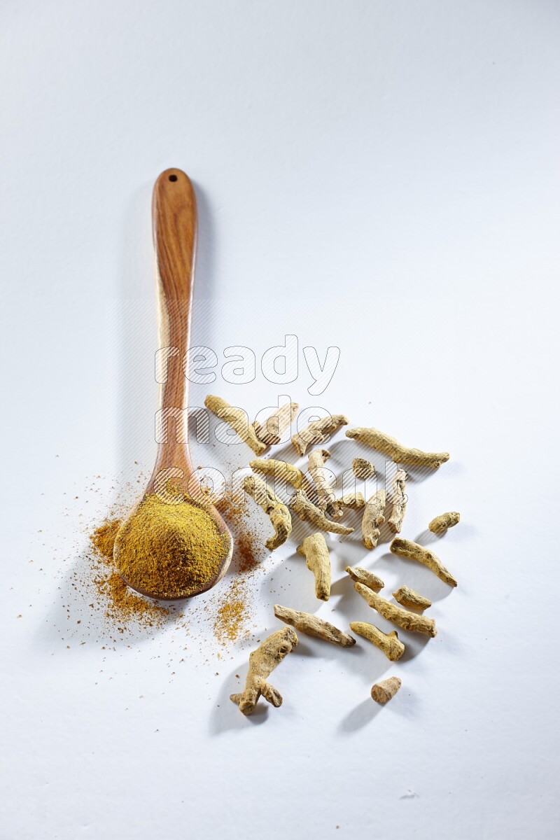 A wooden ladle full of turmeric powder and dried turmeric fingers beside it on white flooring