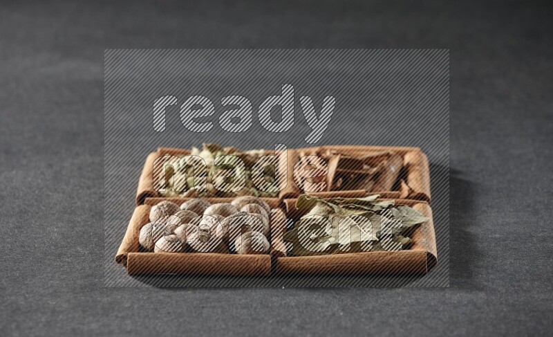 4 مربعات من عيدان القرفة ممتلئة بقطع القرفة وحبوب الهيل وأوراق الغار وجوزة الطيب على أرضية سوداء