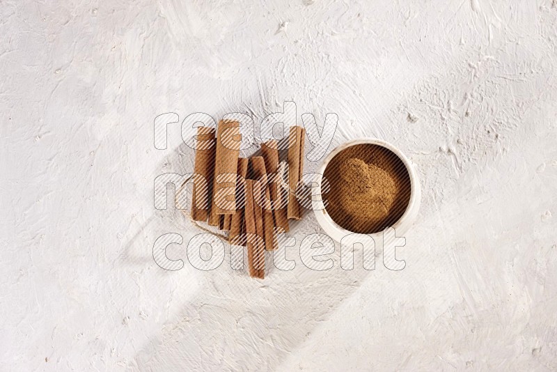 Cinnamon sticks stacked beside a beige bowl full of cinnamon powder on white background