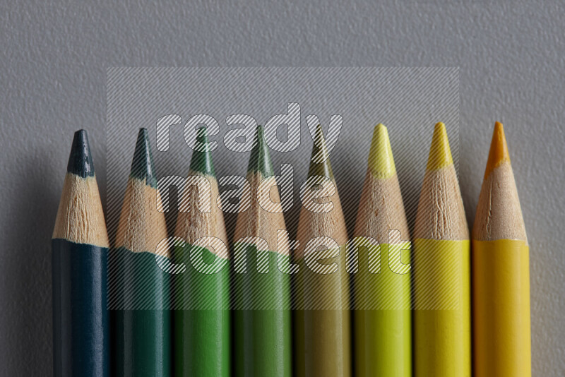 A collection of sharpened colored pencils arranged showcasing a gradient of yellow and green hues on grey background