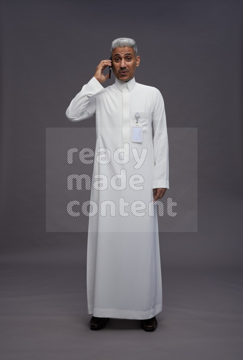 Saudi man wearing thob with pocket employee badge standing talking on phone on gray background