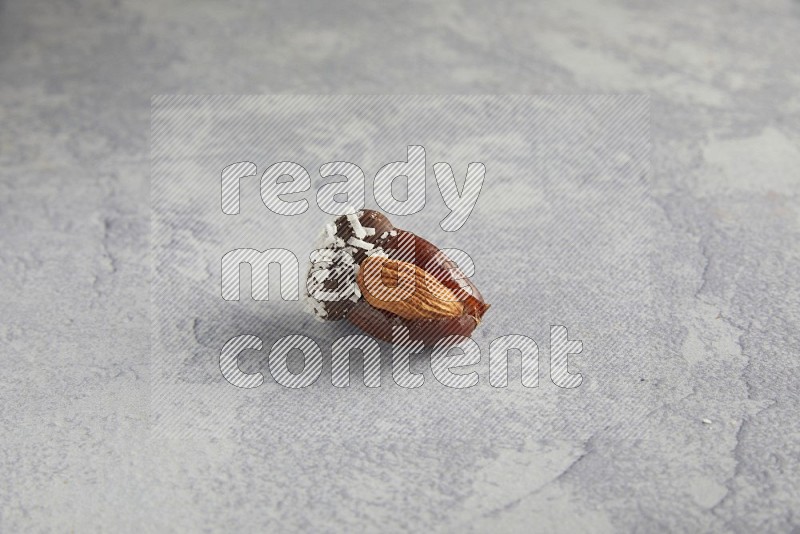 Almond stuffed date covered with dark chocolate and coconut on a light grey background