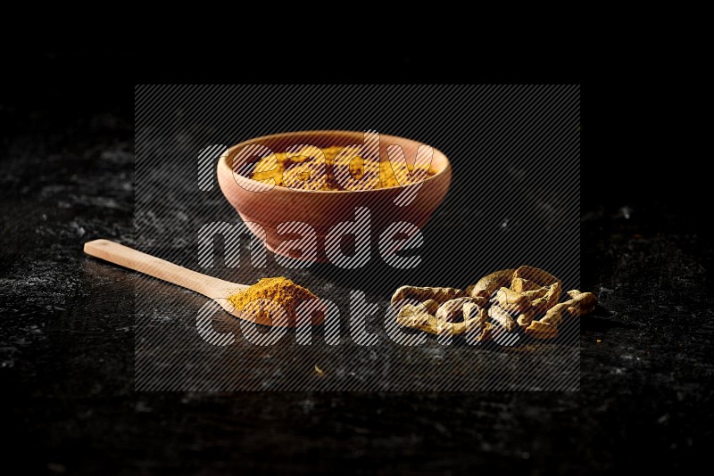 A wooden bowl and a wooden spoon full of turmeric powder with dried turmeric fingers on textured black flooring